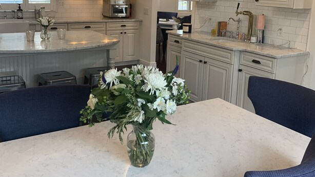 A kitchen with white cabinets, a central island, stainless steel appliances, and blue chairs around a marble table with a vase of flowers. - In Sag Harbor Village. Walk to town, and less than 1/2 mile to Havens Beach. (Sag Harbor)