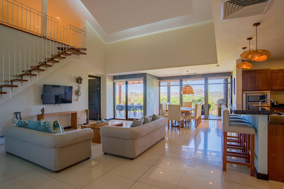 A spacious living area with a high ceiling, a staircase, a flat-screen TV, a sofa, a dining area with a table and chairs, and a kitchen with bar stools.