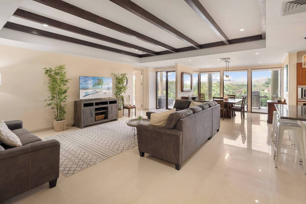A spacious living area with a large gray sofa, a flat-screen TV, and a dining table with chairs. There are potted plants and a patterned rug on the floor.