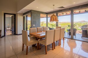 A dining area with a wooden table and chairs, large windows with a view of the outdoors, and a ceiling fan. - Reserva Conchal Resort - Carao Complex (Cabo Velas)