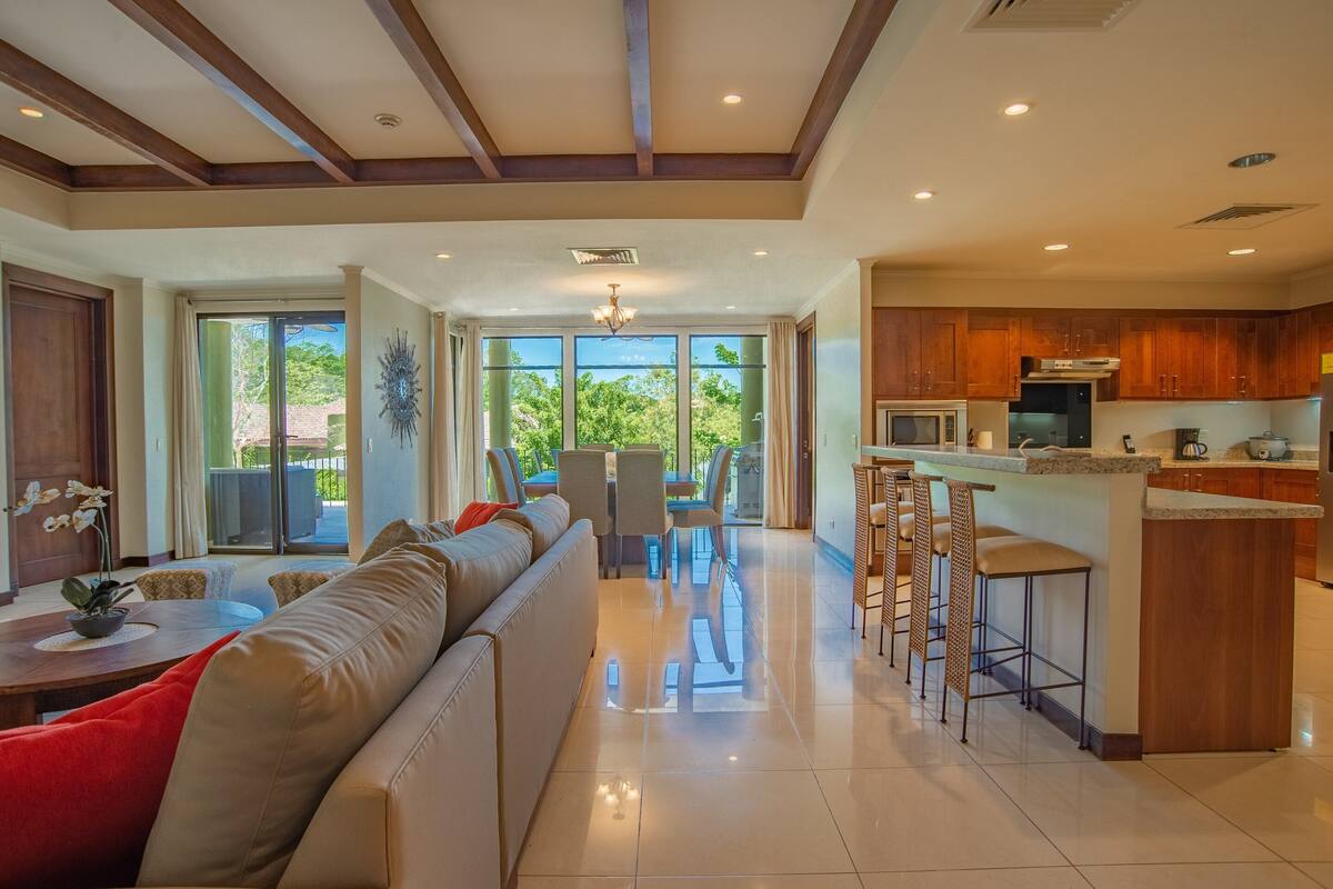 A spacious living area with a large sofa, a dining table, and a kitchen area with wooden cabinets and a bar stool.