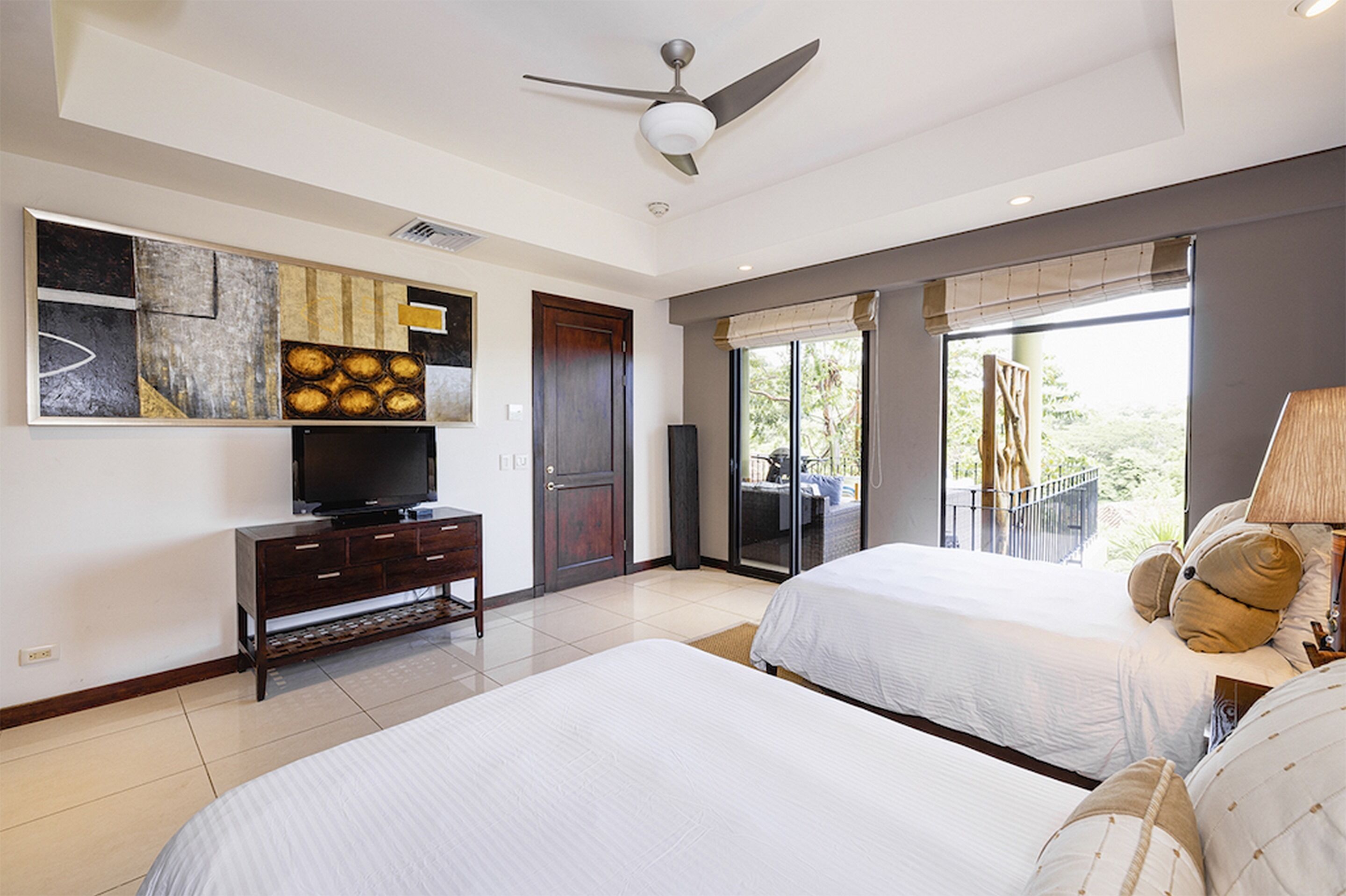 A hotel room with two beds, a TV on a wooden cabinet, a ceiling fan, and a balcony with a view.