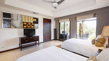 A hotel room with two beds, a TV on a wooden cabinet, a ceiling fan, and a balcony with a view.