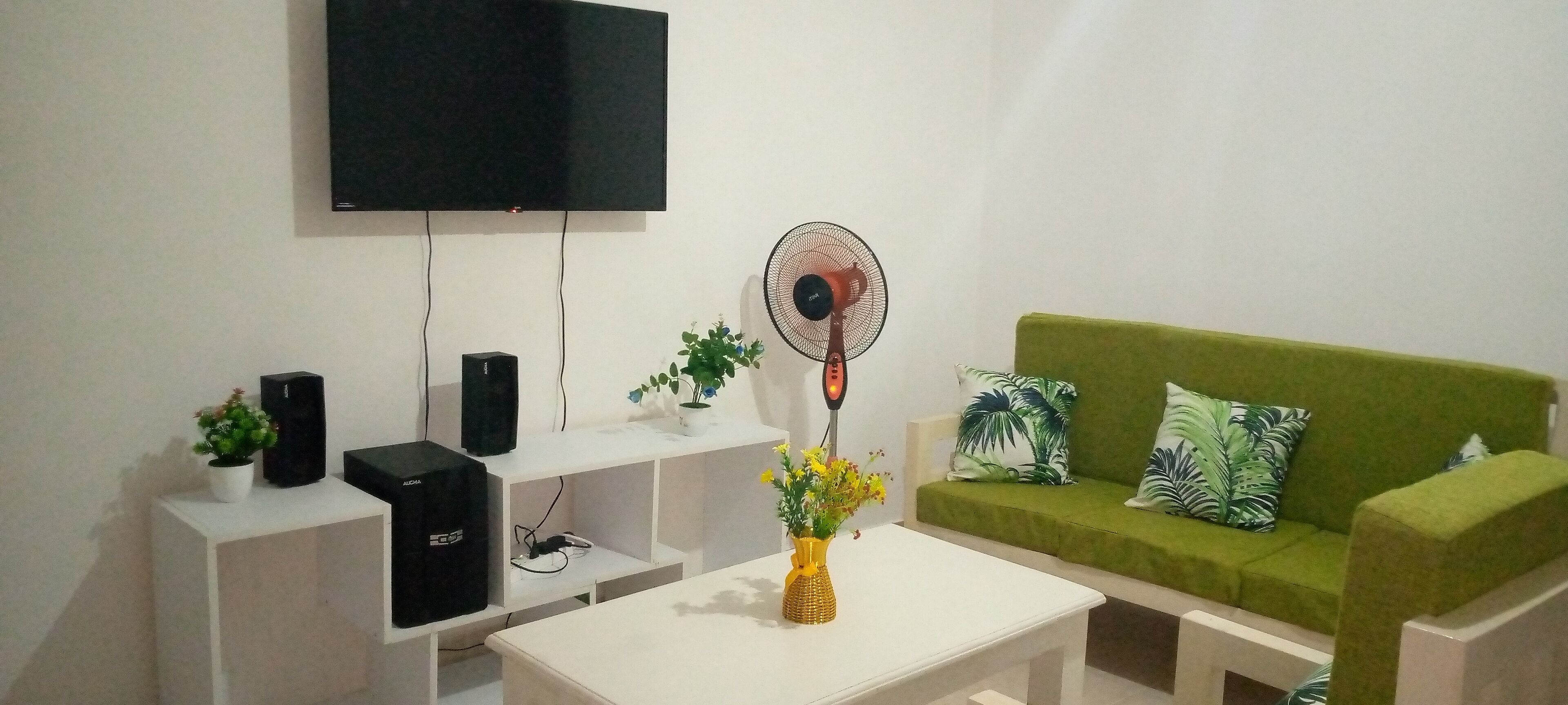 Un salon équipé d’un téléviseur à écran plat, d’un ventilateur sur pied, d’un canapé vert agrémenté de coussins à motifs, d’une table basse blanche et d’une petite plante posée sur une table d’appoint blanche.
