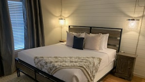 Een net opgemaakt bed met een houten hoofdbord, een witte deken en meerdere kussens. Twee nachtkastjes met lampen, een houten bijzettafeltje en een raam met zonwering.