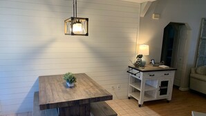 Een eetgedeelte met een houten tafel, banken, een hanglamp, een kleine plant in pot en een witte kast met lades.