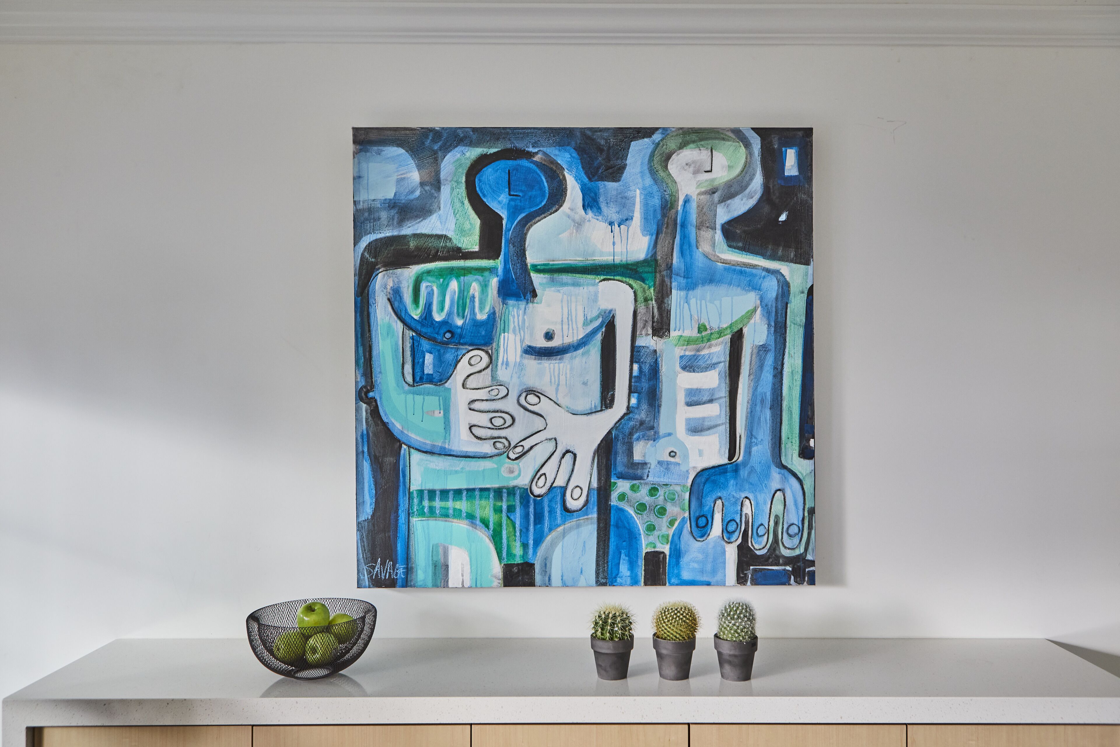 Una pintura de formas abstractas y un cuenco de manzanas verdes sobre una superficie blanca.