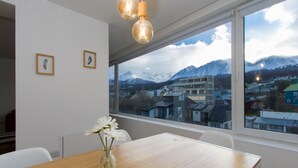 Una habitación con una mesa de madera, sillas blancas, un jarrón con flores, cuadros en la pared y un amplio ventanal con vistas a la montaña.