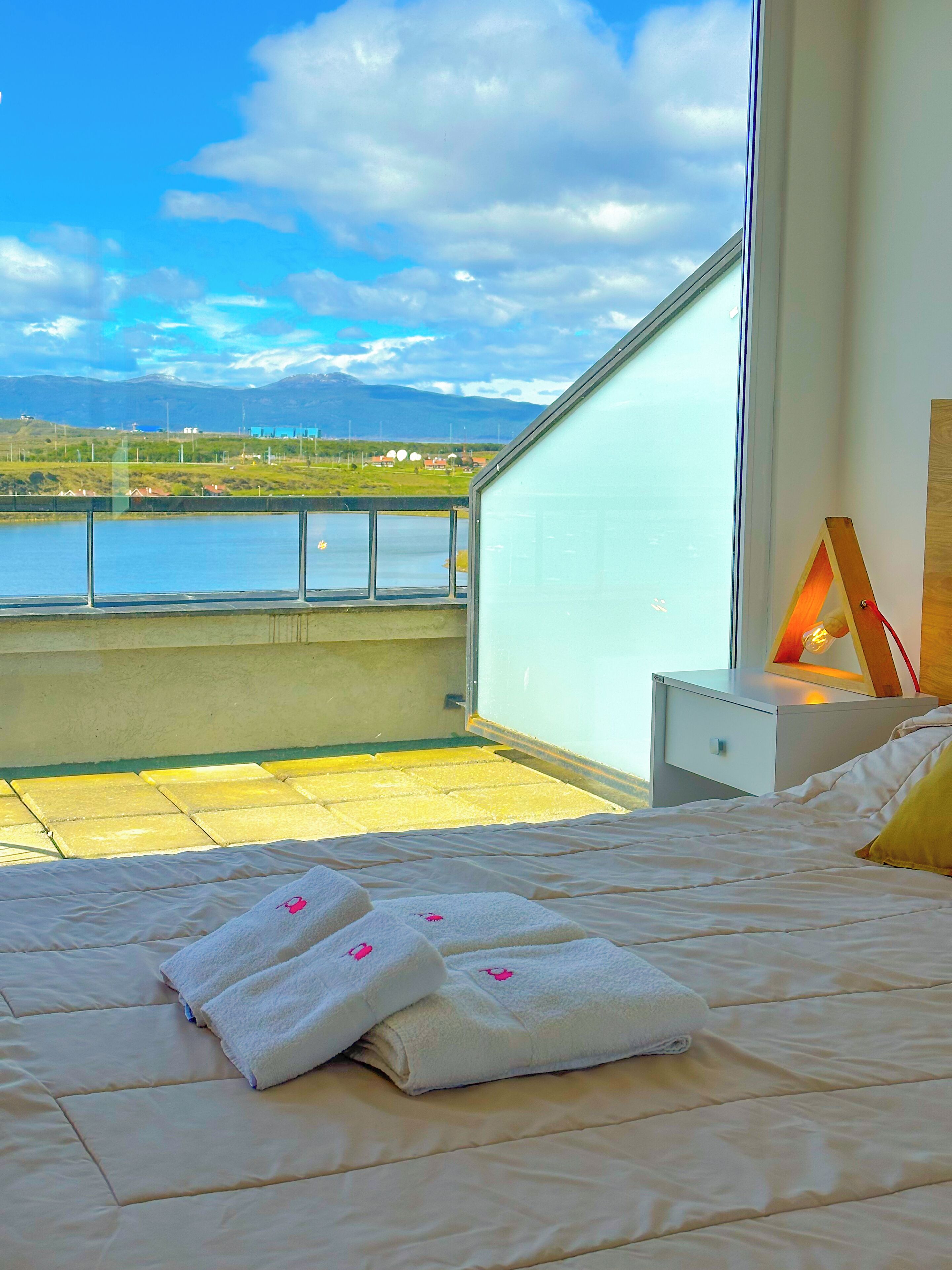 A bedroom with a large window overlooking a body of water and a view of distant hills.
