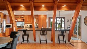 A modern kitchen with wooden cabinets, a central island with bar stools, and a dining area with a round table and chairs. - The Great Escape - Picturesque Sebago Lake (Raymond)
