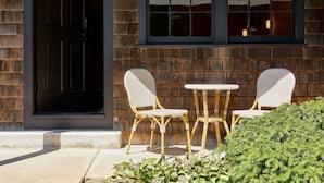 A building entrance with a dark door, a brick wall, and a window with a view of trees. There are two wicker chairs and a small round table outside. - Bluebird Dennisport (Dennis Port)