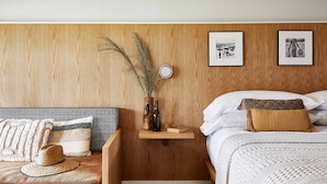 A bedroom with a wooden headboard, a bedside table with a hat and a bottle, and two framed pictures on the wall. - Bluebird Dennisport (Dennis Port)