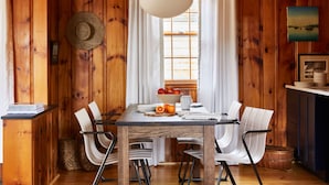 A wooden dining room with a table set for four, white chairs, a hanging light fixture, and a window with curtains. - Bluebird Dennisport (Dennis Port)
