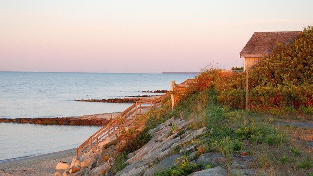 A coastal scene with a wooden staircase leading down to the beach, surrounded by rocks and vegetation. - Bluebird Dennisport (Dennis Port)