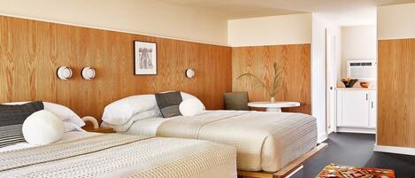 A modern bedroom with two beds, a small table, and a view into the kitchen.