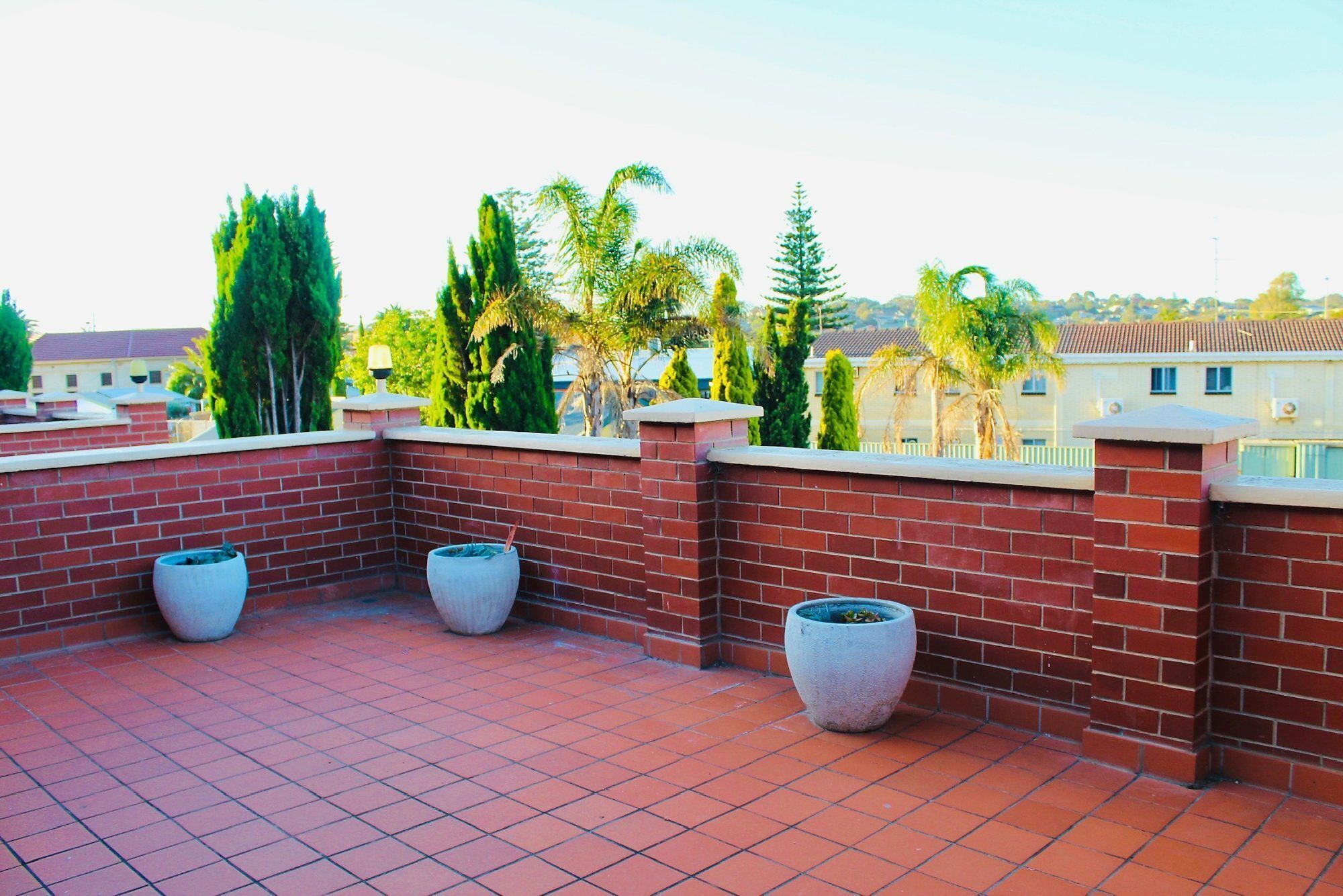 A rooftop terrace with red brick walls, three large planters, and a view of surrounding buildings and trees.