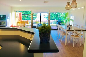 A modern kitchen with a black countertop, a potted plant, and a dining area with white chairs and a table. - Prime Location – Heart of Victor Harbor: Steps away from the Beach, Top Floor, Ocean View, Wi-Fi, Smart TV (Victor Harbor)