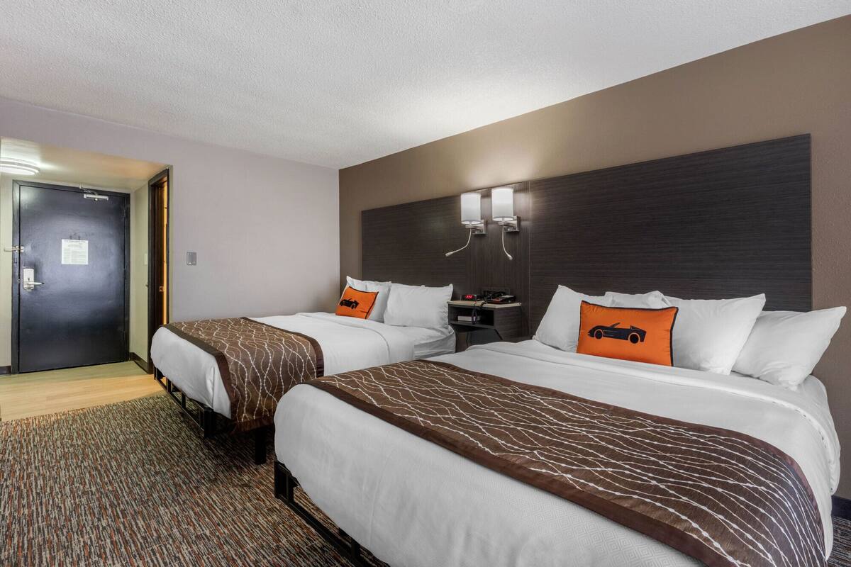 Hotelkamer met twee bedden, een donker hoofdbord, een nachtkastje met een lamp en een deur met een bordje.