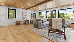 Ein geräumiges Wohnzimmer mit Holzboden, einem grauen Sofa, einer Holzkονstruktion an der Decke und großen Fenstern mit Blick auf Bäume.