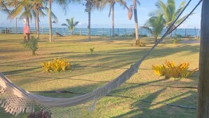 A hammock under a wooden ceiling, a wooden bench with a small plant, and a view of a grassy area with palm trees and a fence. - Pousada Vila Rosa (Porto Seguro)