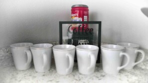 A coffee-themed display with a red can of coffee and a framed sign, surrounded by six white mugs on a marble countertop. - Keizer Valley Estate I 3BR Family Stay Near Salem Oregon (Keizer)