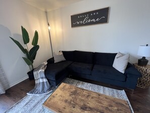 Ein Wohnzimmer mit einem dunkelblauen Sofa, einem Holztisch, einer Topfpflanze und einem Wandschild mit der Aufschrift 'HELLO AND welcome'.