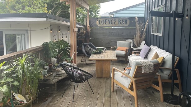 A covered outdoor seating area with wooden furniture, potted plants, and a sign reading "LOUNGE BAR". - Walk to town and beach with beautiful views (Raglan)