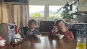 A dining area with a wooden table, two children eating, a decorative mailbox, and a view of mountains through large windows. - Walk to town and beach with beautiful views (Raglan)