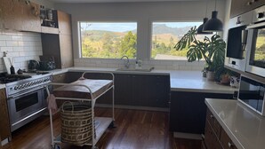 A modern kitchen with a large window, wooden cabinets, stainless steel appliances, and a view of the countryside. - Walk to town and beach with beautiful views (Raglan)