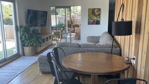 A modern living room with a wooden floor, a round dining table with chairs, a sofa, a TV, and a large window. - Walk to town and beach with beautiful views (Raglan)