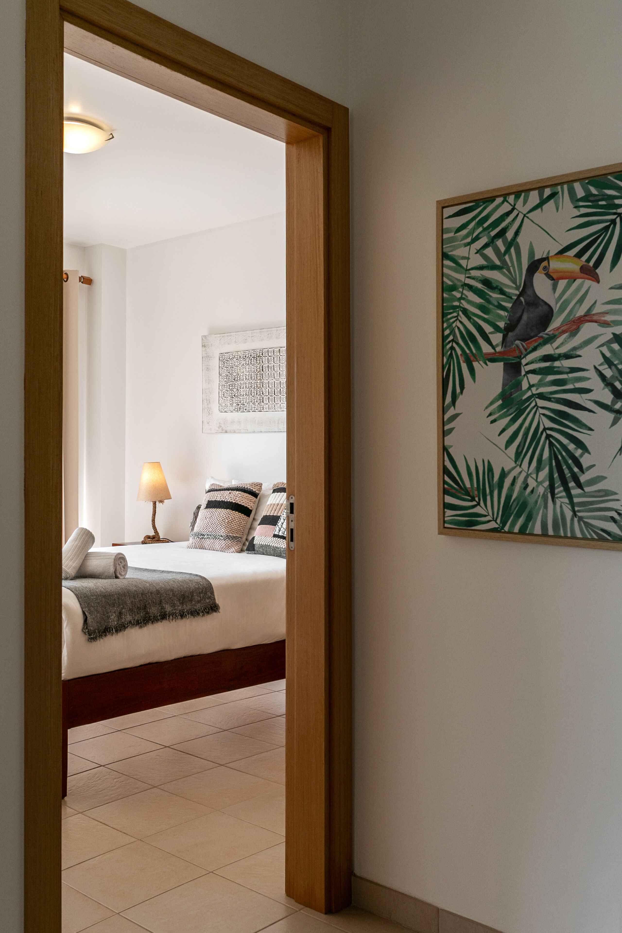 A hotel room with a bed, a nightstand with a lamp, a wall with a tropical bird painting, and a wooden door frame.
