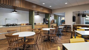 A modern dining area with wooden tables and chairs, a kitchen in the background, and pendant lighting. - Fairfield by Marriott Inn & Suites Phoenix West Chandler (Phoenix)