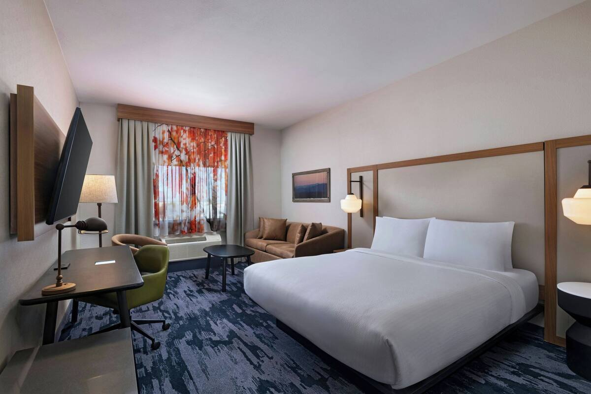 A hotel room with a large bed, a desk with a lamp, a chair, a small table, and a television.