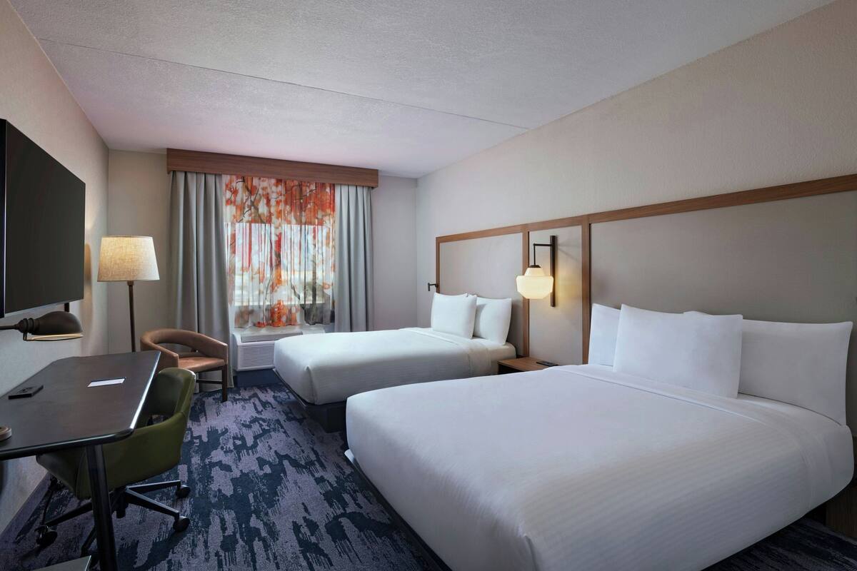 A hotel room with two beds, a desk, a chair, a TV, and a window with curtains.
