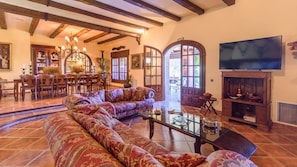 A spacious living room with a large sectional sofa, a glass coffee table, and a television on a wooden stand. There is a dining area with a wooden table and chairs, and a kitchen area with a wooden cabinet and a stove.