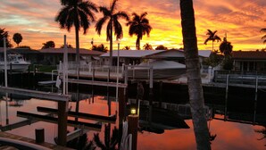 Een jachthaven met aangemeerde boten, een overdekt gedeelte en palmbomen tegen een zonsondergang.