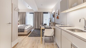 Ein modernes Hotelzimmer mit einem Bett, einer Essecke mit Tisch und Stühlen, einer Küche mit Spüle und Herd sowie einem großen Fenster mit Vorhängen.
