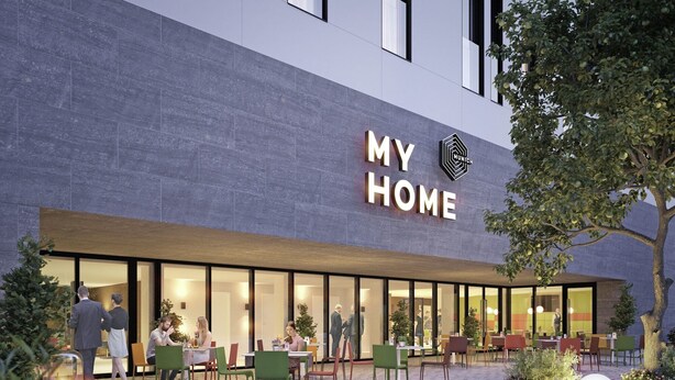 Ein modernes Gebäude mit einem Schild, auf dem "MY HOME" steht, und einer Terrassenfläche mit Außenbestuhlung.