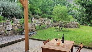 A patio with a stone wall, a wooden table with a bottle and glasses, and a small garden area with a tree and a bench. - A cozy nest for a pair (Civitella Paganico)