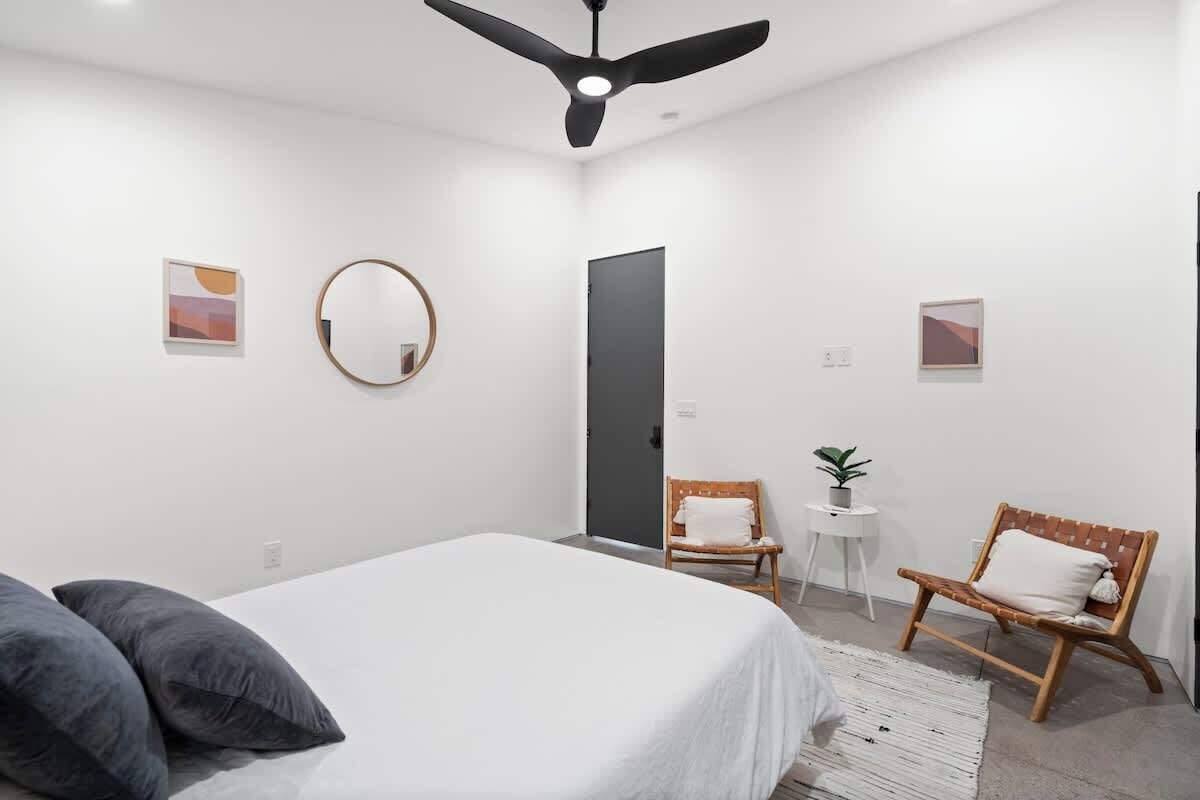 A modern bedroom with a bed, two chairs, a round mirror, and a ceiling fan.