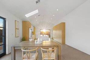 A modern dining area with a long table, chairs, and a view of the sunset through a window.