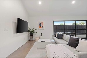 A modern living room with a flat-screen TV, a sofa, a coffee table, and a large window. - Modern Home w/ Heated Pool (Phoenix)