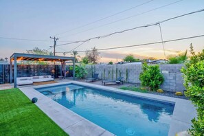 A swimming pool with a covered seating area, surrounded by a low wall and greenery.