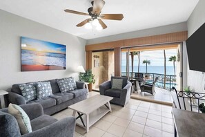 A living room with a sofa, armchairs, a coffee table, and a ceiling fan. There is a painting of a sunset on the wall.