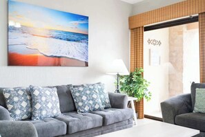 A living room with a grey sofa, patterned cushions, a painting of a beach scene, a lamp, and a plant.