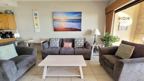 A living room with a sofa set, a coffee table, a painting of a sunset, and a wall-mounted sign.