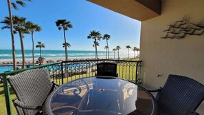 A balcony with a glass table and chairs, overlooking a beach with palm trees and a pool.