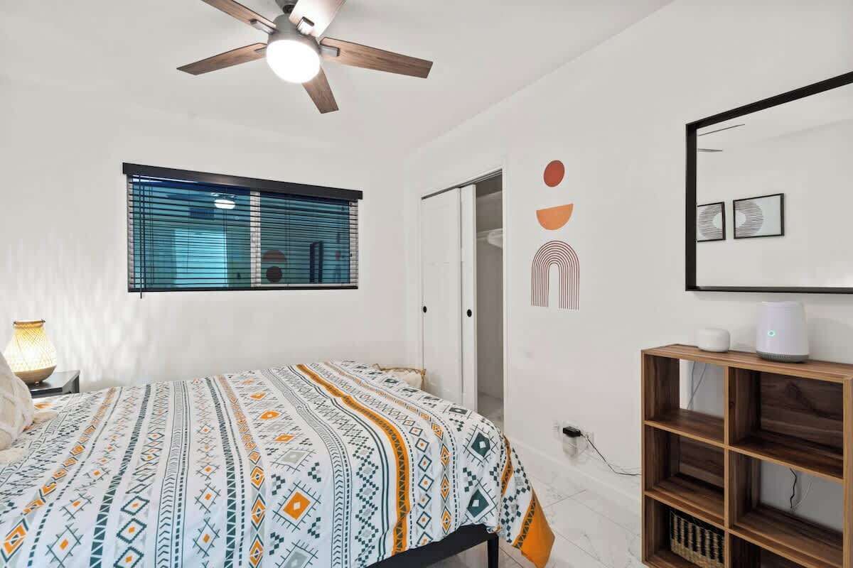 Een slaapkamer met een bed, een plafondventilator, een raam met zonwering, een nachtkastje met lamp en een houten plank.