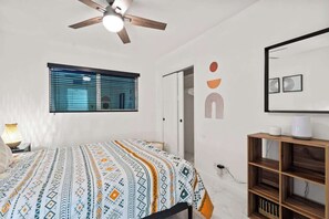 A bedroom with a bed, a ceiling fan, a window with blinds, a bedside table with a lamp, and a wooden shelf.