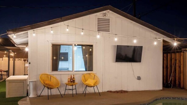 Una casa con piscina, dos sillas amarillas, una mesita con botellas y luces de cadena.
